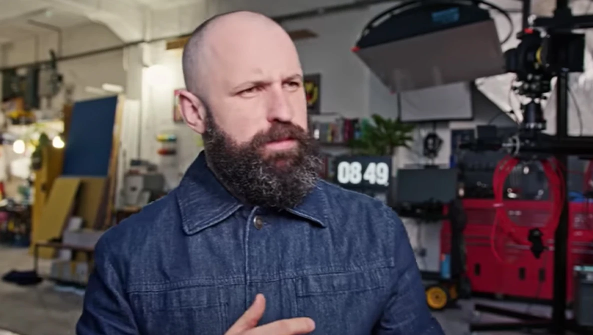 Bearded man in denim shirt speaking in professional studio with camera equipment visible