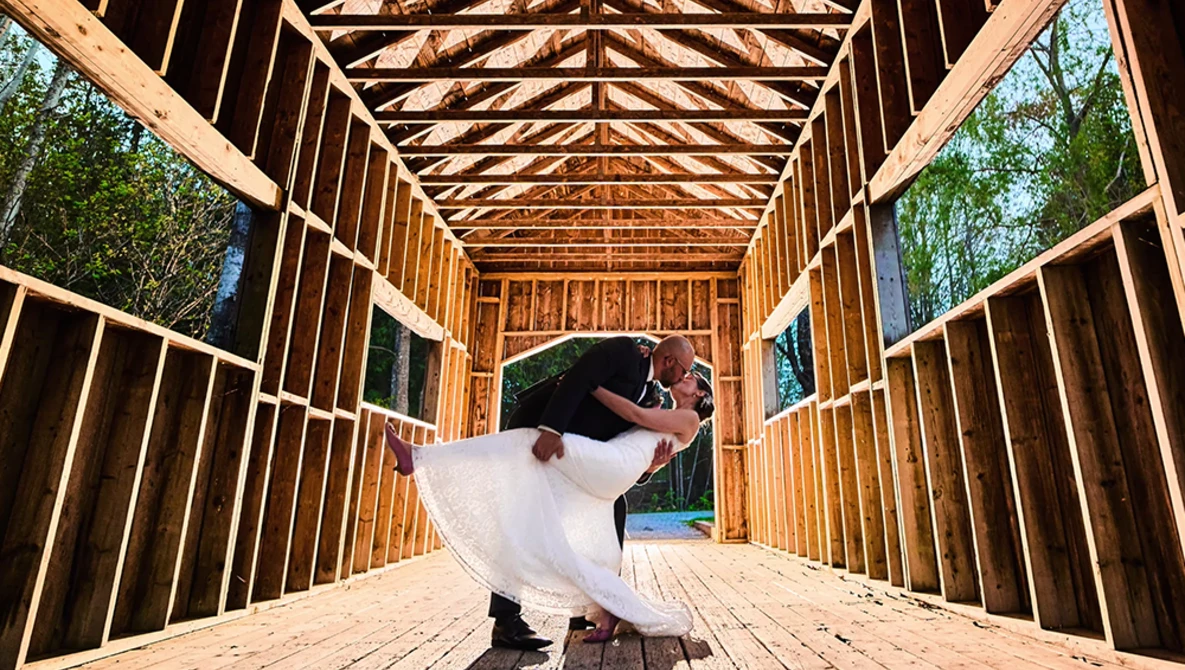 Bride in white dress twirling inside wooden barn structure with open sides and geometric roof framing.