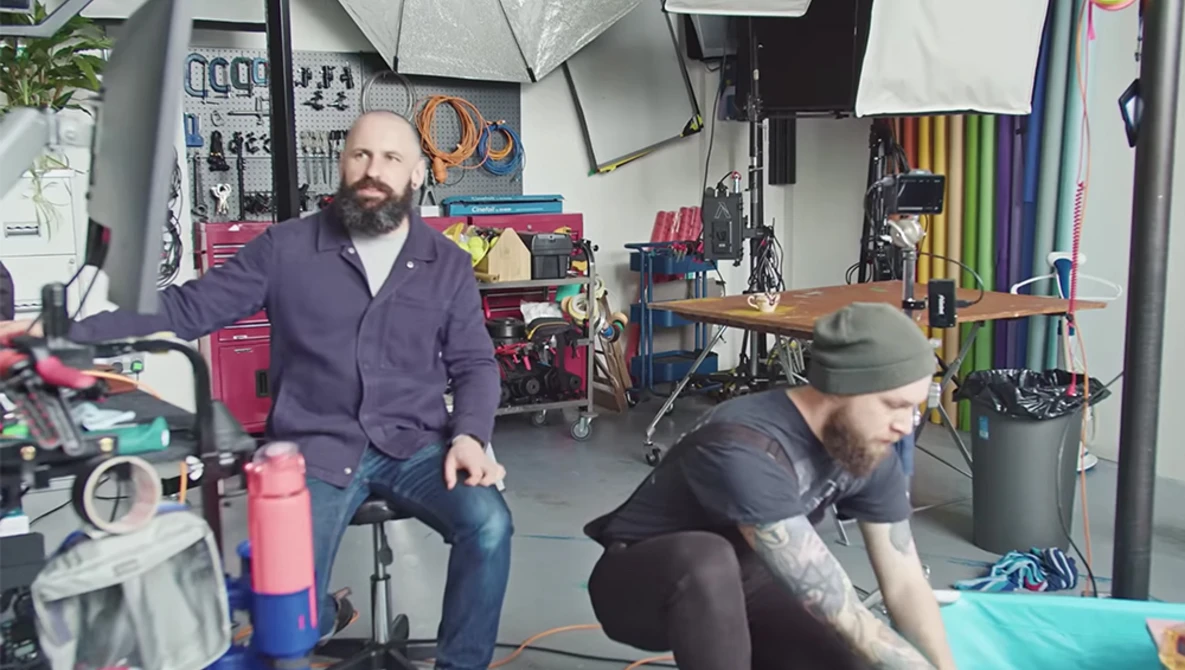 Two people in a workshop setting surrounded by photography and creative equipment on shelves and walls.