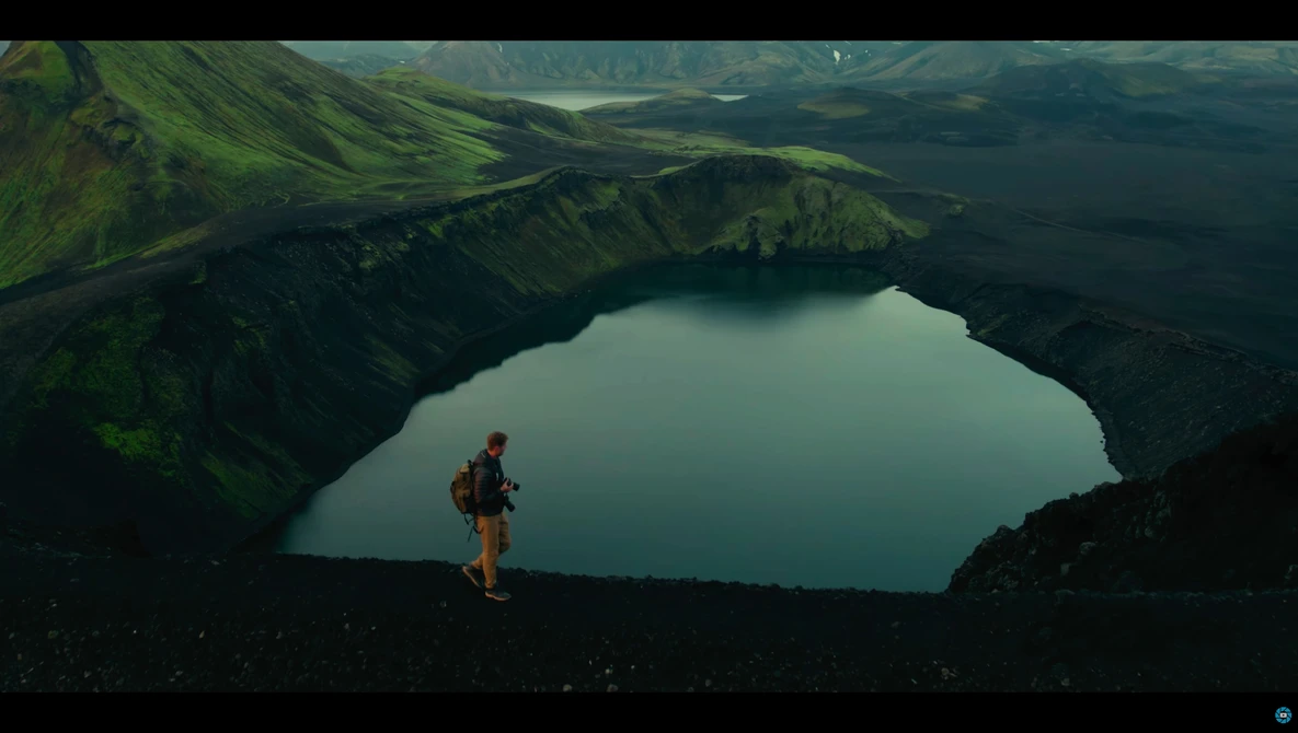 Hiker in red jacket standing on dark volcanic terrain overlooking a turquoise crater lake surrounded by green mountains.