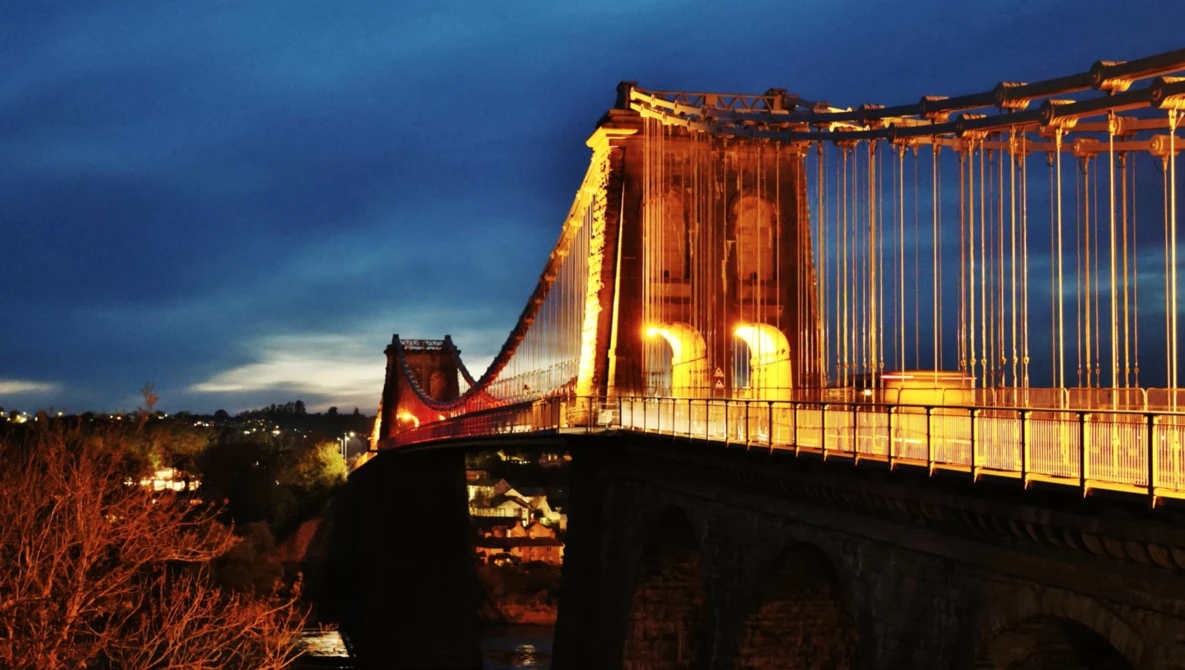 Suspension bridge illuminated in golden light at dusk against a dramatic blue sky.