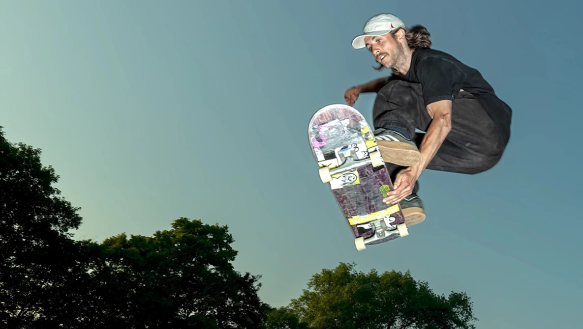 Skateboarder airborne against clear sky with trees below, wearing white cap and black clothing.