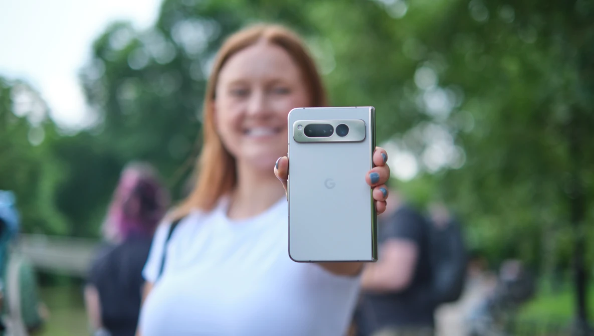 Woman holding a smartphone with dual rear cameras toward the camera in an outdoor park setting.