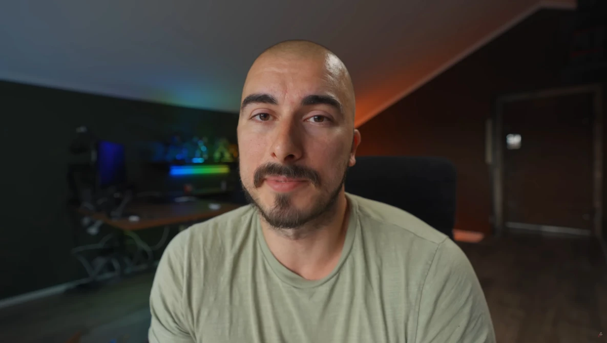 Man with shaved head and goatee wearing light gray v-neck t-shirt in dimly lit room.