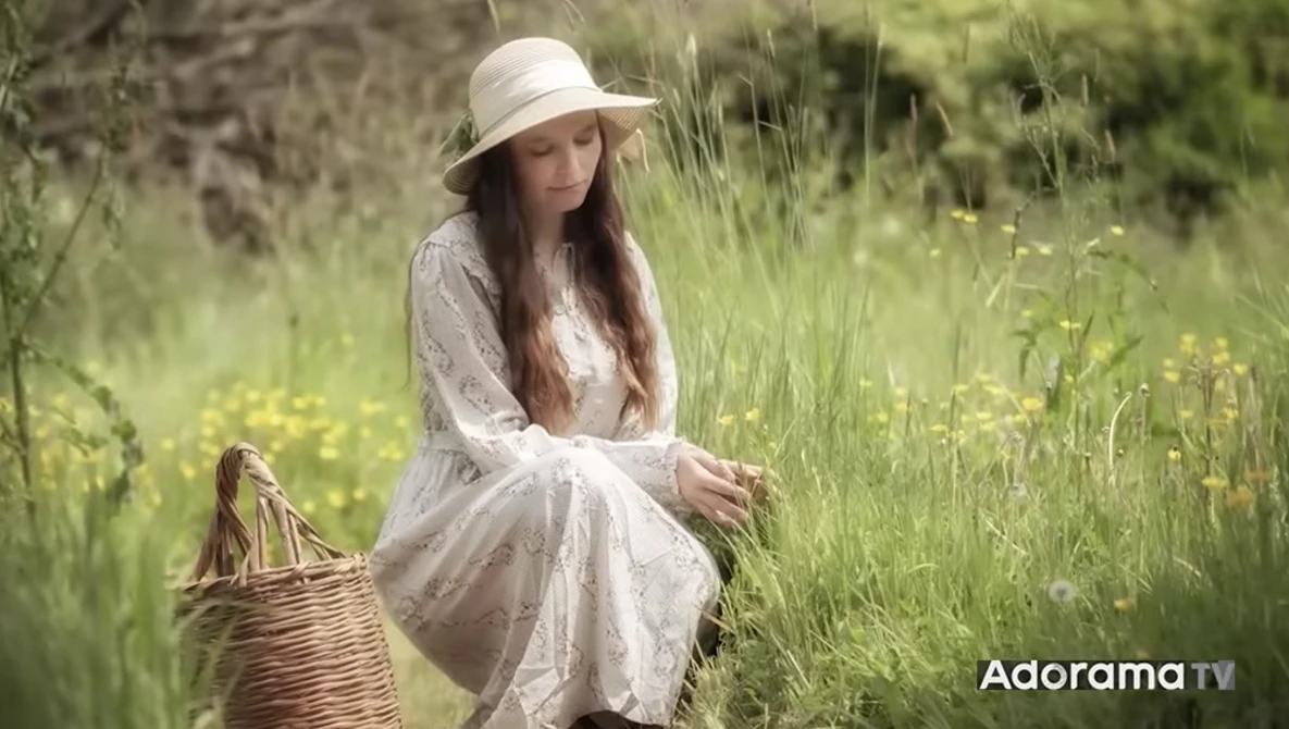 Woman in white dress and hat crouching in wildflower meadow beside wicker basket.