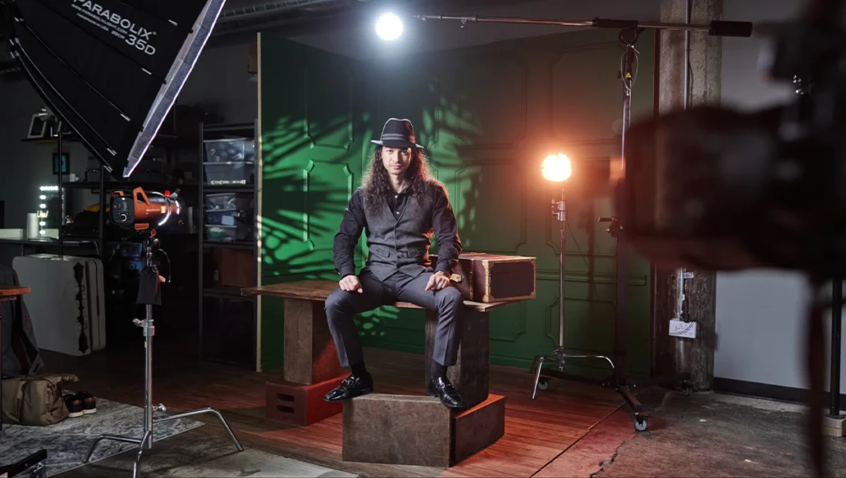 Photographer sitting on wooden box in studio with professional lighting setup and green backdrop.