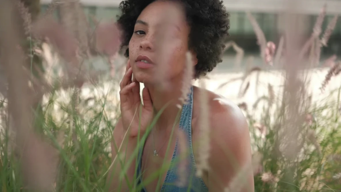 Person with curly hair among tall grasses, hand to face in contemplative pose.