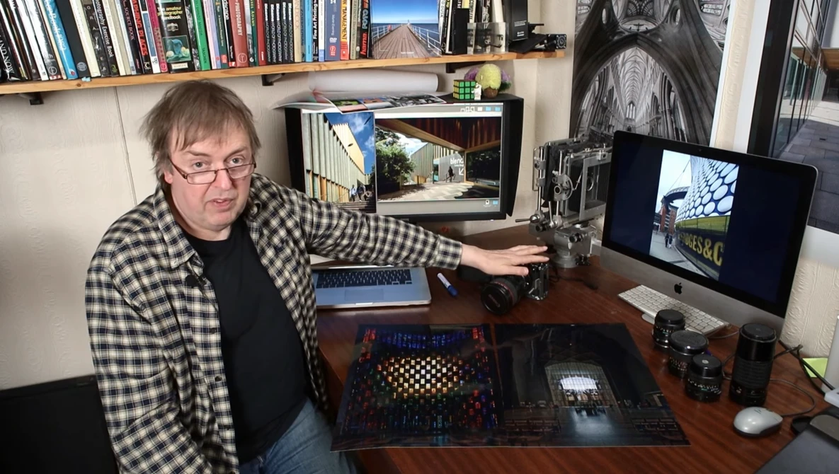 Photographer at desk reviewing images on monitor with camera equipment and shelving visible.