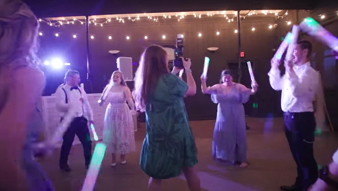 Wedding guests dancing under purple and warm string lights at an indoor reception venue.