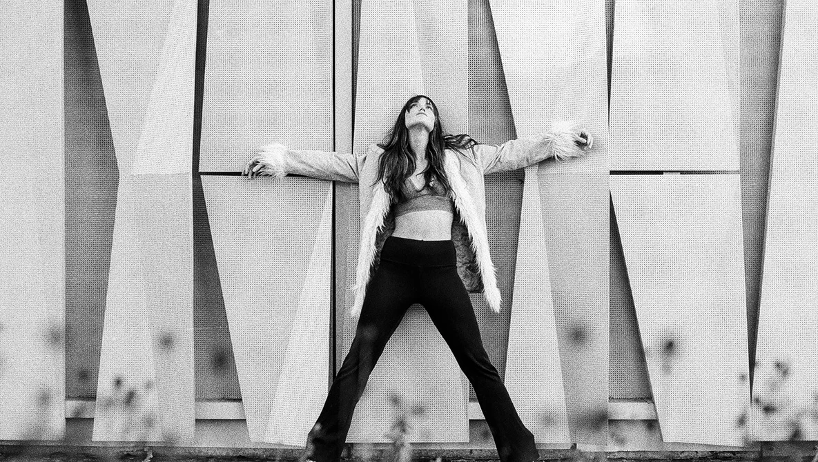 Woman in crop top and black pants striking a wide stance against geometric concrete wall panels.