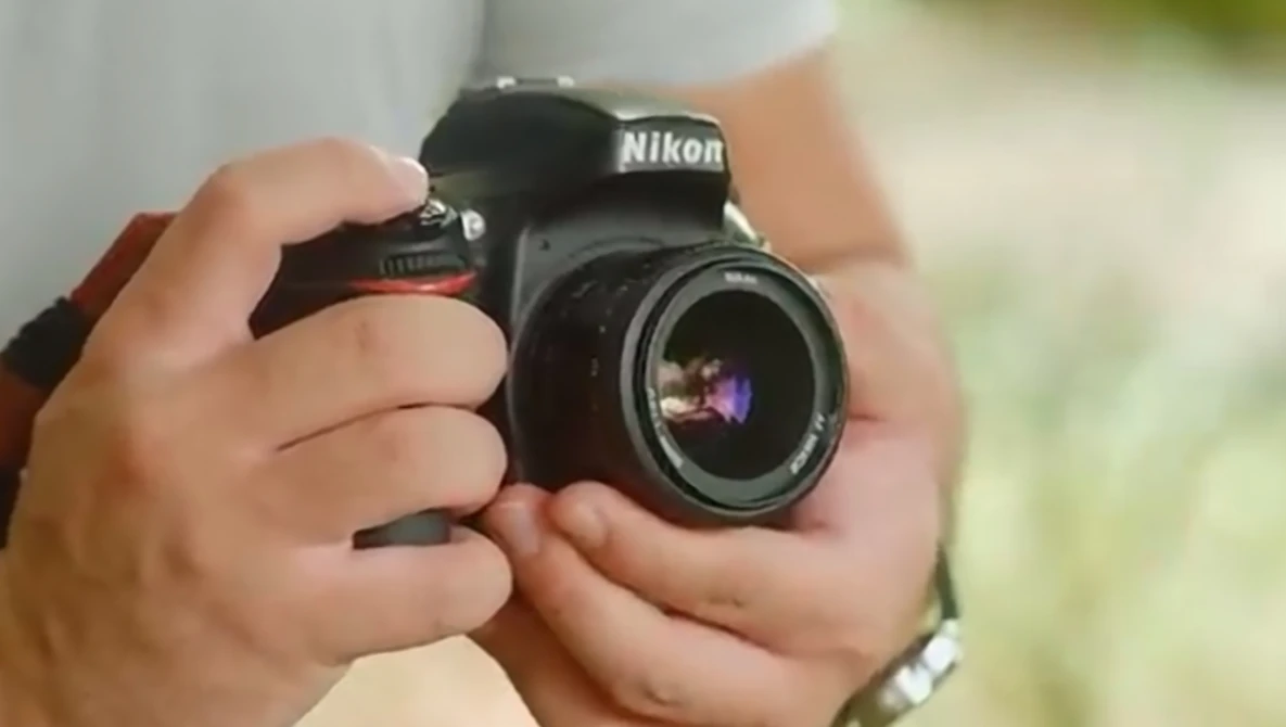 Hands holding a Nikon DSLR camera with a wide-angle lens against a blurred green background.