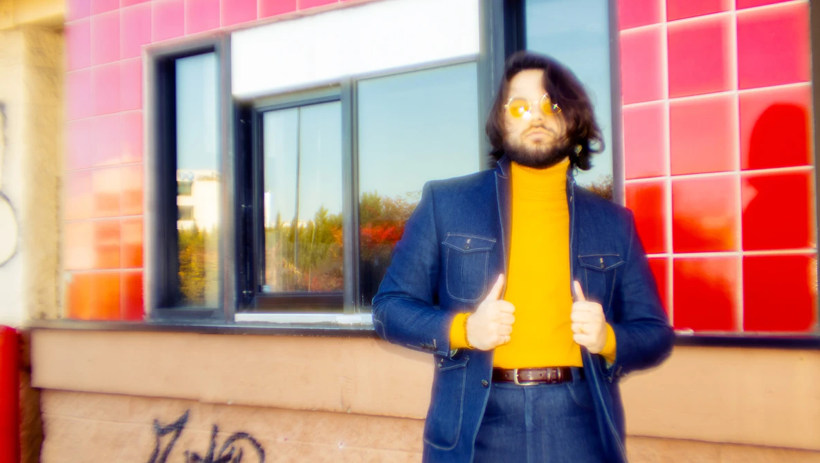 Person in denim jacket and yellow shirt standing against pink tiled wall with windows.