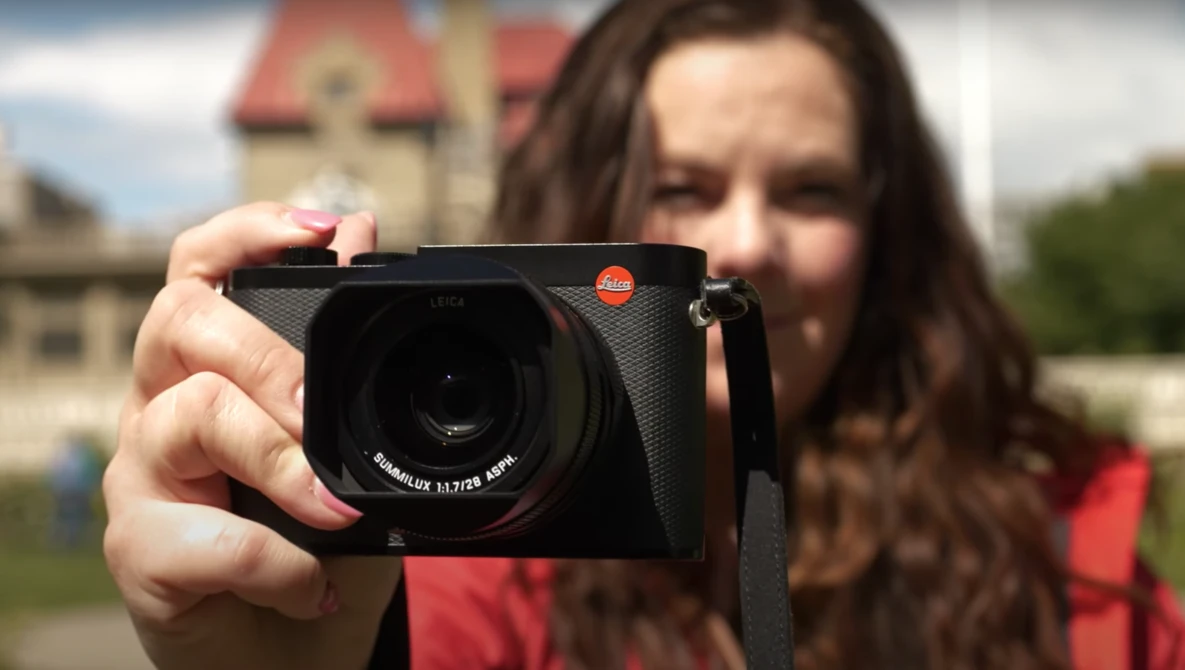 Woman holding a Leica camera toward the viewer, with a historic building blurred in the background.