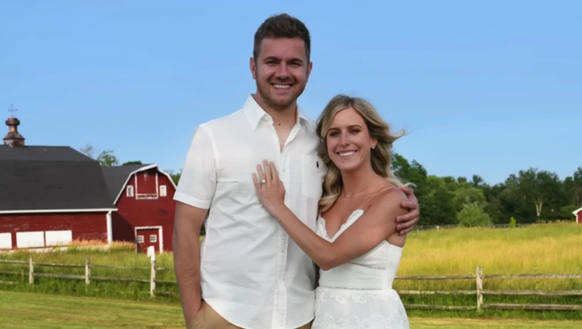 Couple in white clothing posing together in front of a red barn and green pasture.