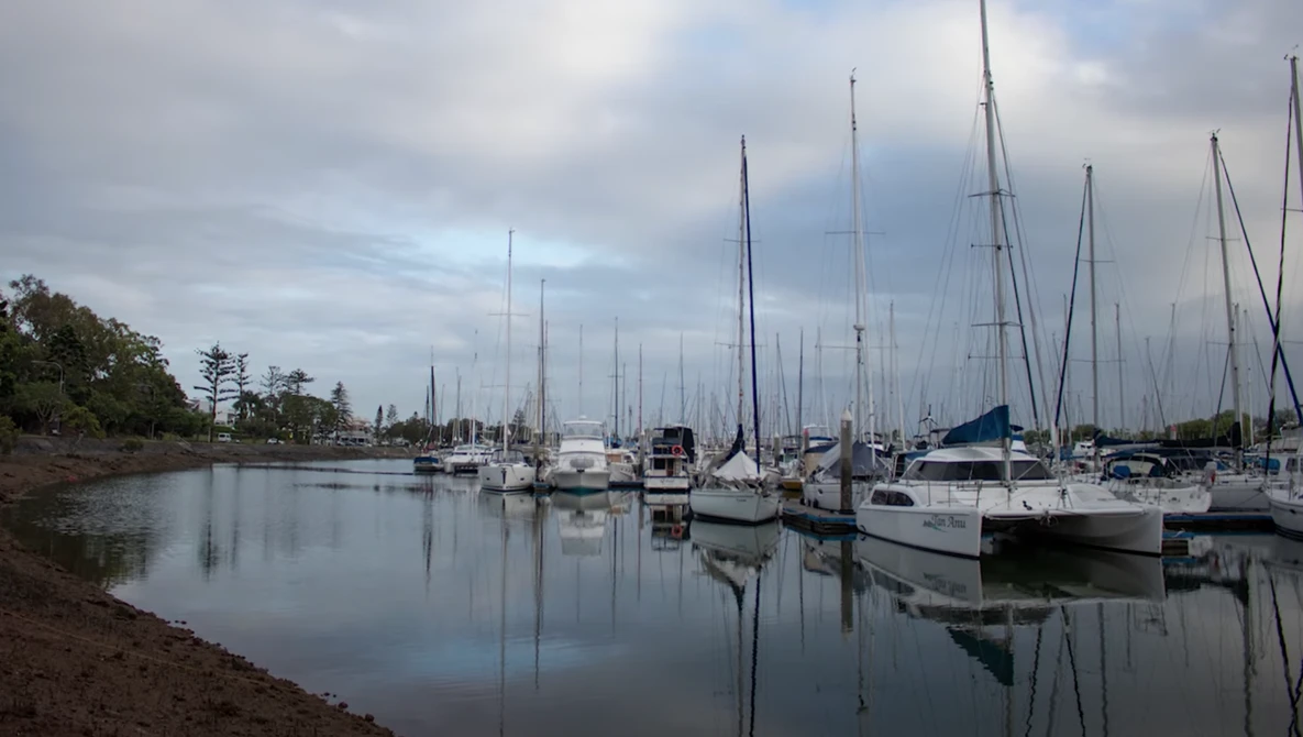 Sailboats moored in calm harbor water with perfect mirror reflections under overcast sky.