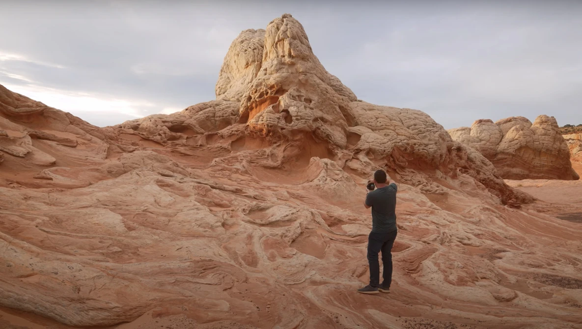 Photographer in dark clothing standing on layered sandstone formations, framing a tall rock pinnacle with camera.
