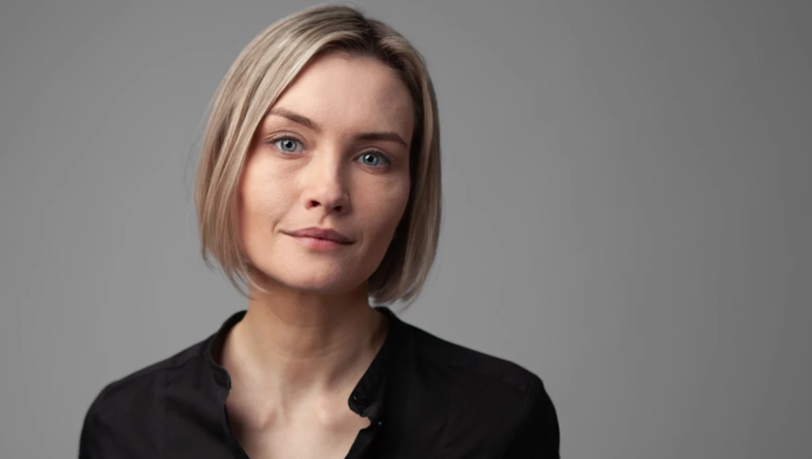 Woman with shoulder-length blonde hair wearing a black shirt against a neutral gray background.