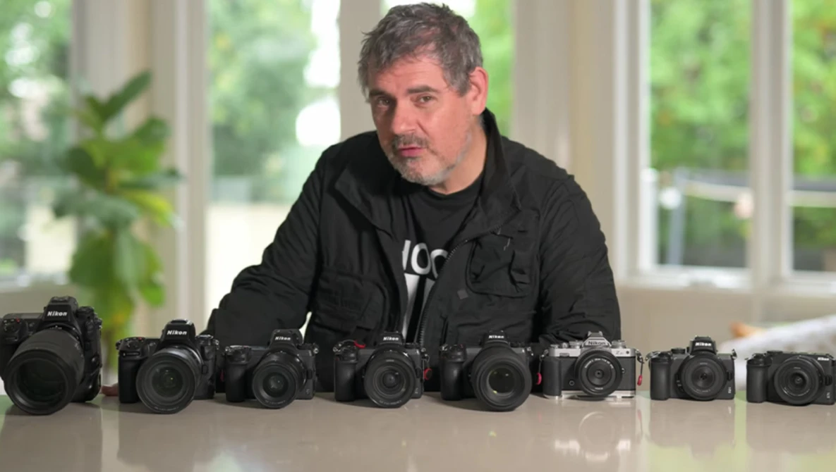 Photographer standing behind nine Nikon DSLR cameras arranged in a line on a white table.