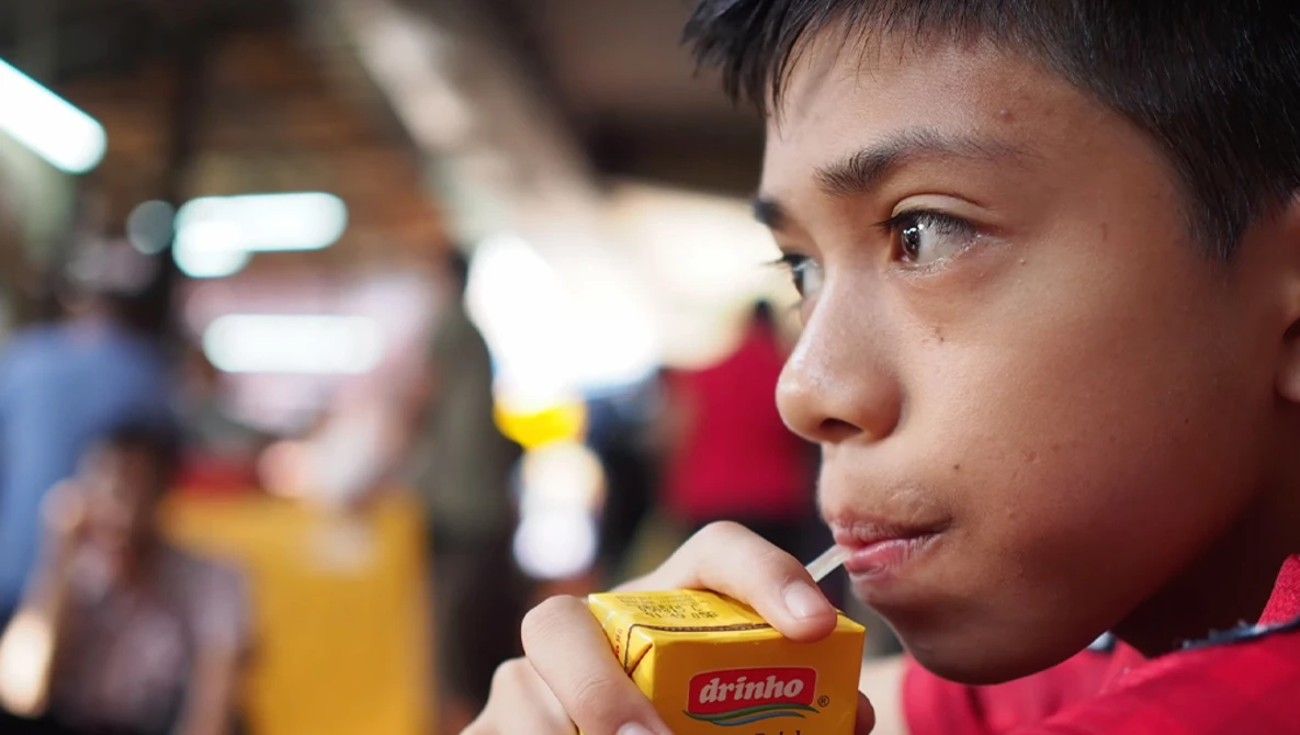 Young person in red shirt holding a yellow beverage box in a crowded indoor space.