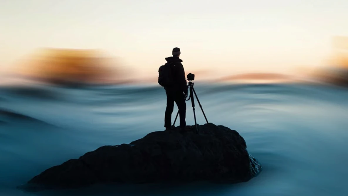 Silhouetted photographer standing on a rocky outcrop with tripod-mounted camera during golden hour sunset.