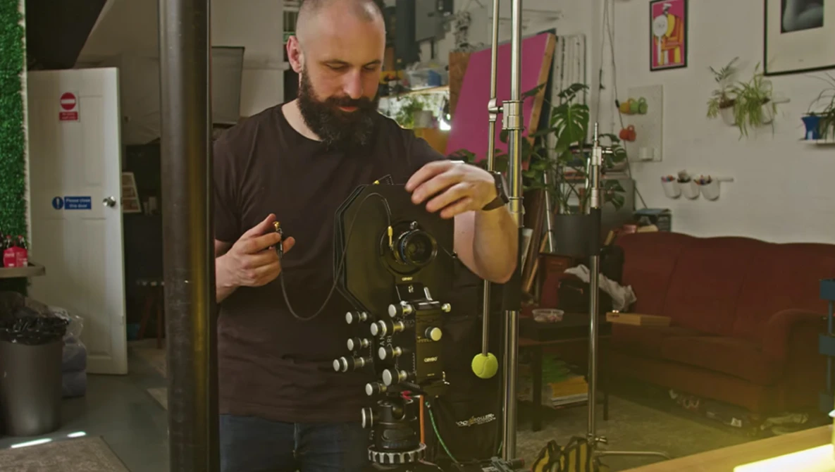 Bearded man examining a complex mechanical device with brass components and hanging weights in a studio setting.