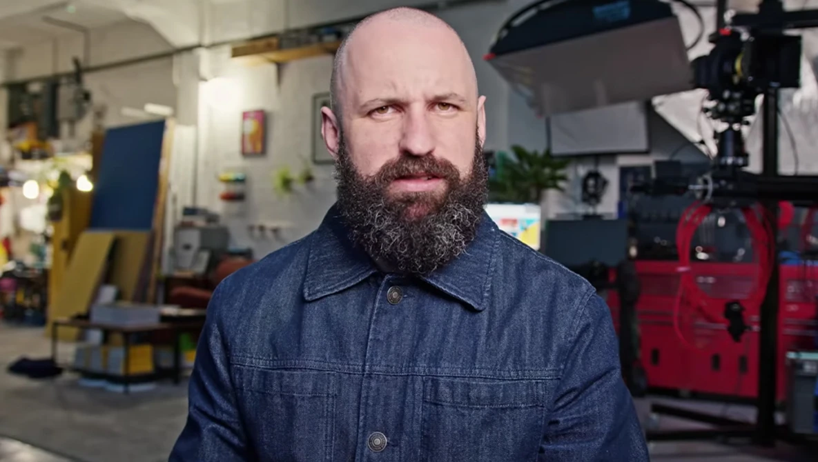 Man with full beard wearing denim shirt in workshop studio setting.