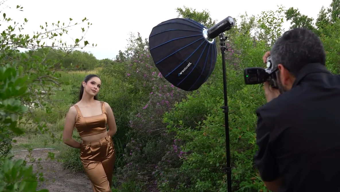 Photographer shooting a woman in bronze-toned clothing outdoors using a large parabolic softbox reflector.