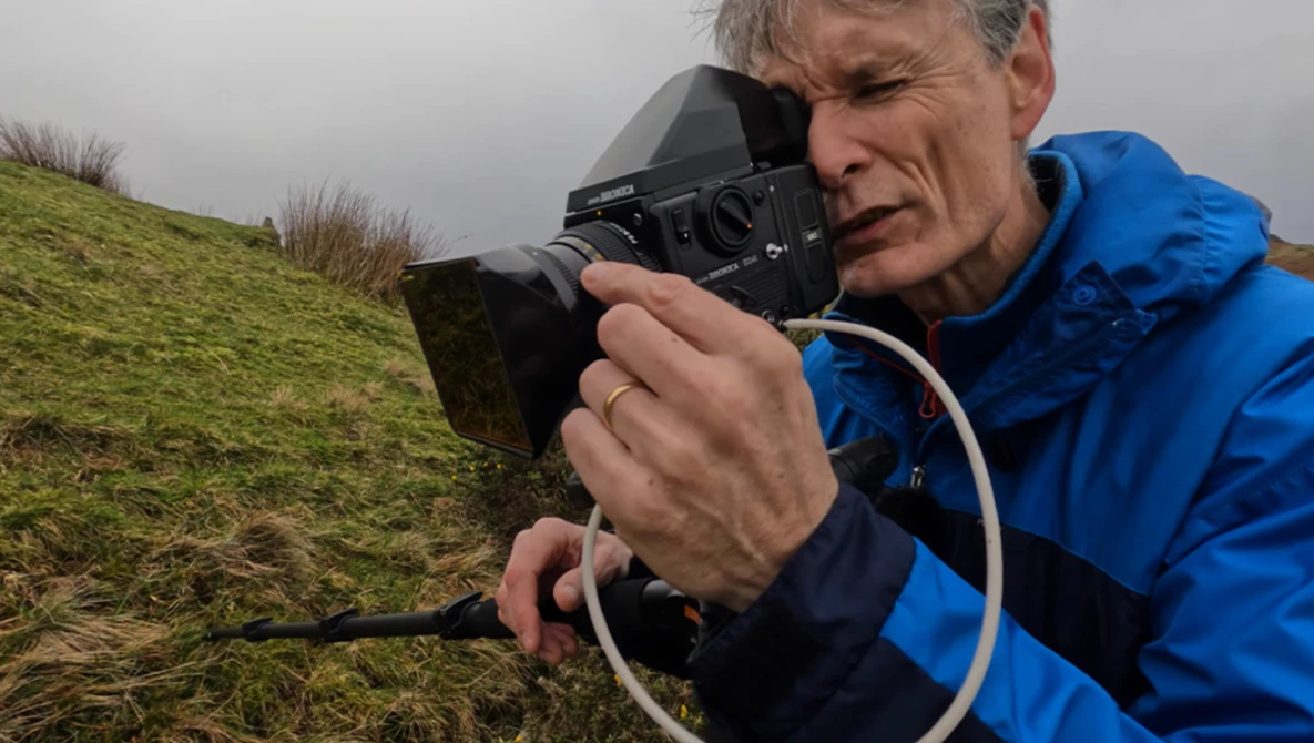 Photographer in blue jacket holding film camera to eye while composing shot on hillside.
