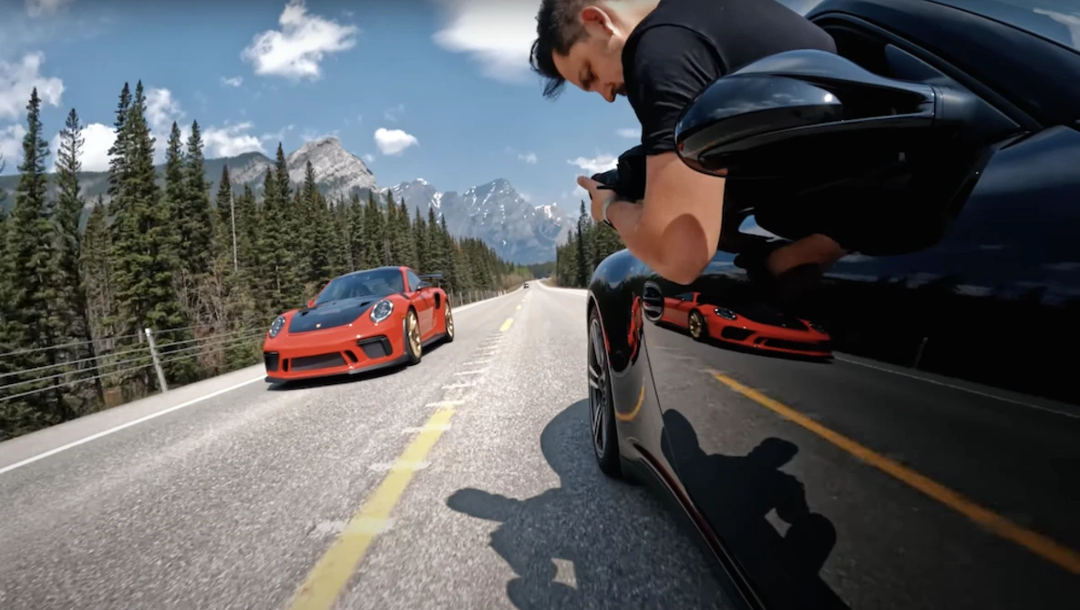 Two sports cars driving on a mountain highway with coniferous forest and peaks in the background.