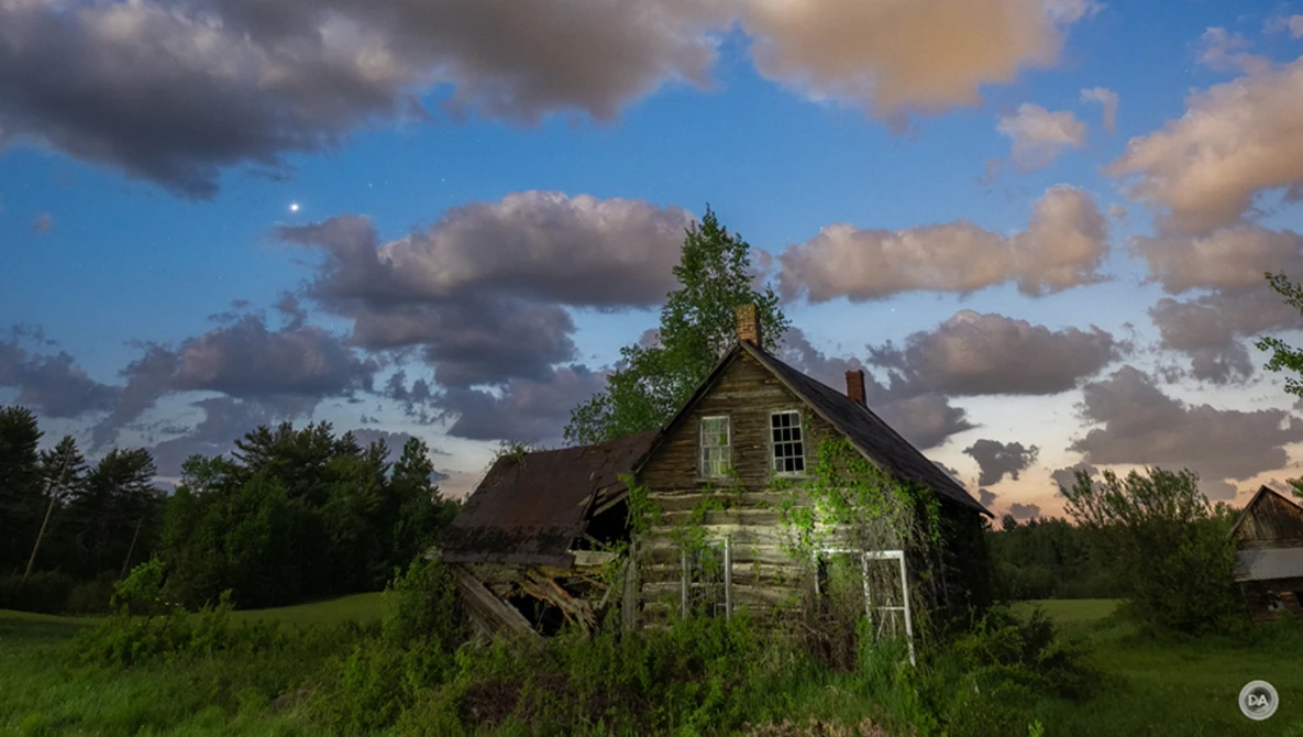 Abandoned wooden house surrounded by overgrown vegetation under a dramatic cloudy sky.