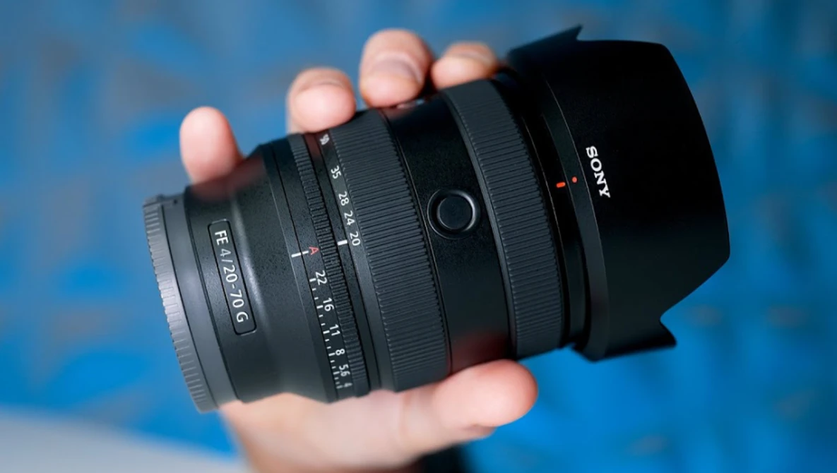 Hand holding a Sony camera lens with black barrel and hood against blue background.