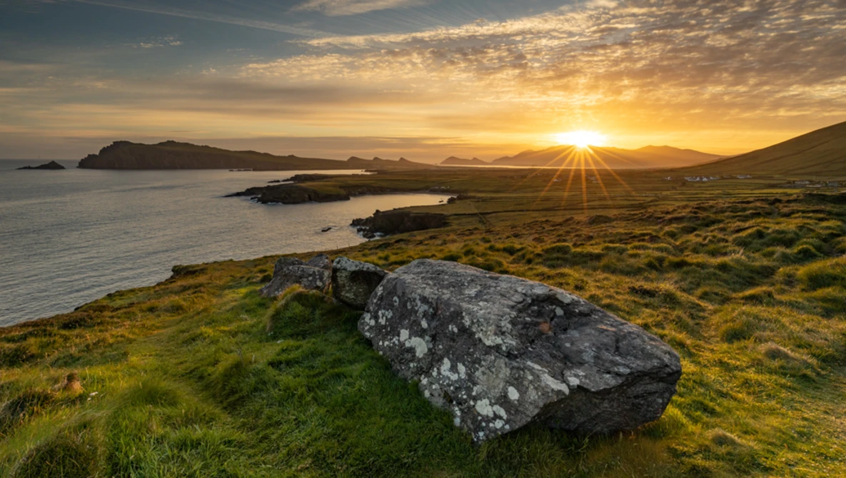 Lichen-covered boulder on grassy coastal hillside at sunset, with distant islands and mountains across the water.