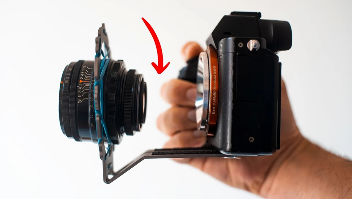 Camera lens mounted on a mirrorless camera body held in a hand, with a red arrow indicating the lens attachment point.
