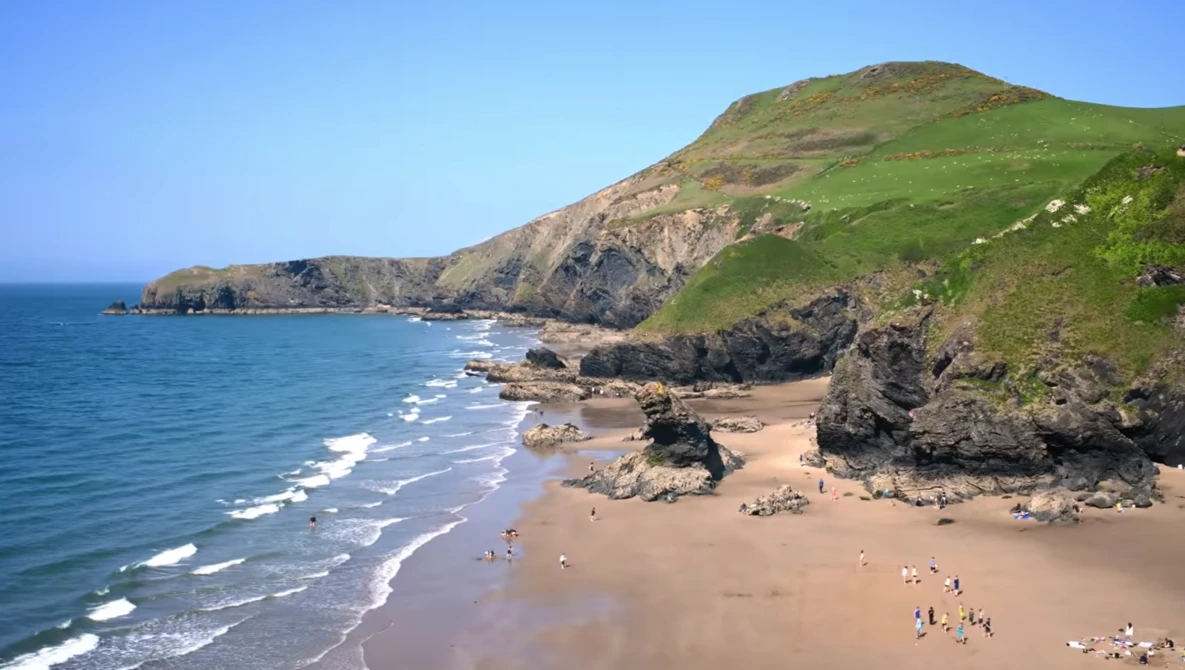 Sandy beach backed by steep green cliffs with rocky outcrops and ocean waves rolling toward shore.