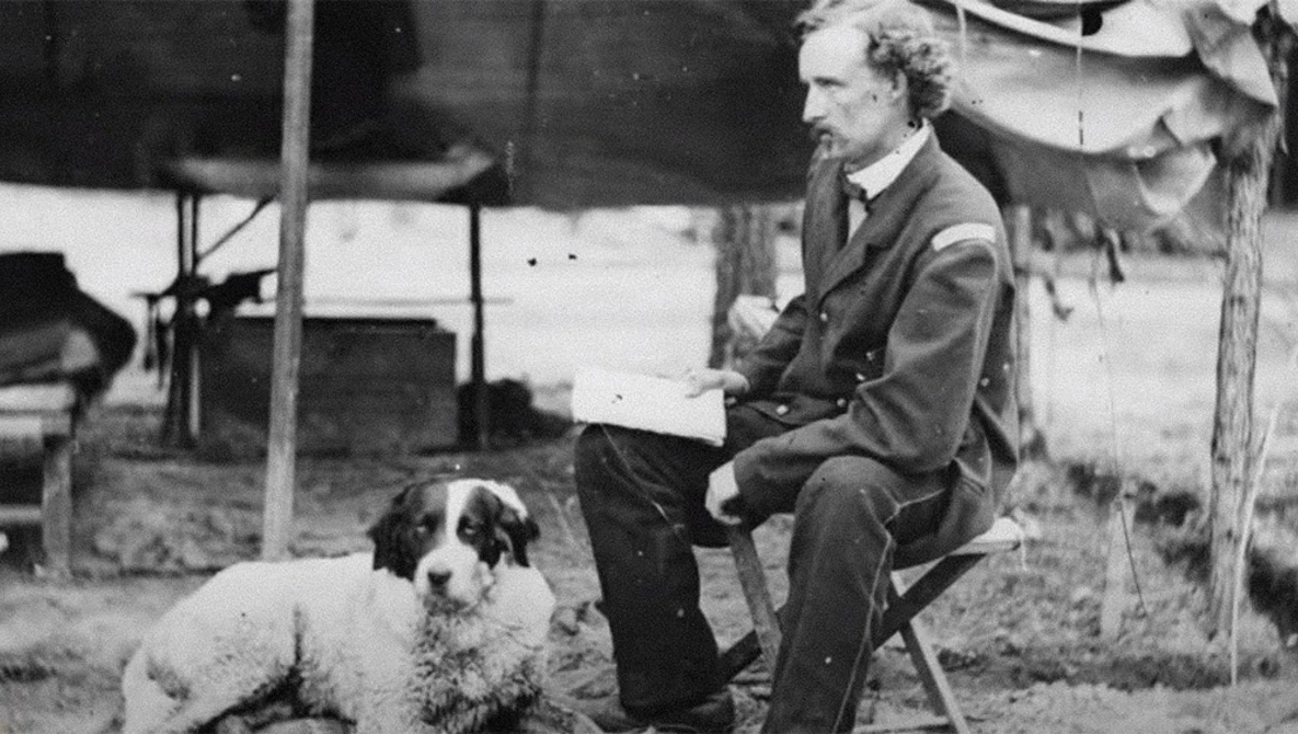Man in suit seated outdoors holding a document, with a seated dog beside him.