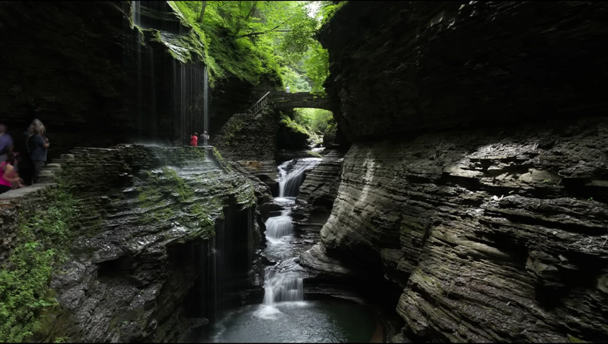 Narrow gorge with cascading water flowing between steep layered rock walls and lush green canopy above.
