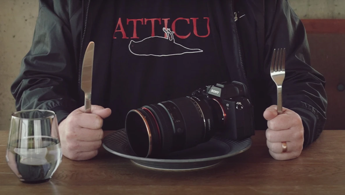 Photographer holding utensils and camera in dining setup, wearing ATTICU branded shirt.