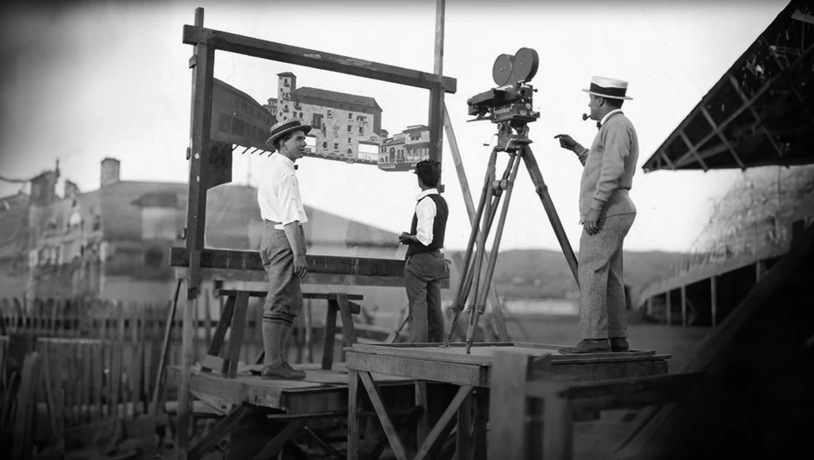 Film crew operating a movie camera on a platform overlooking a European city during early cinema production.