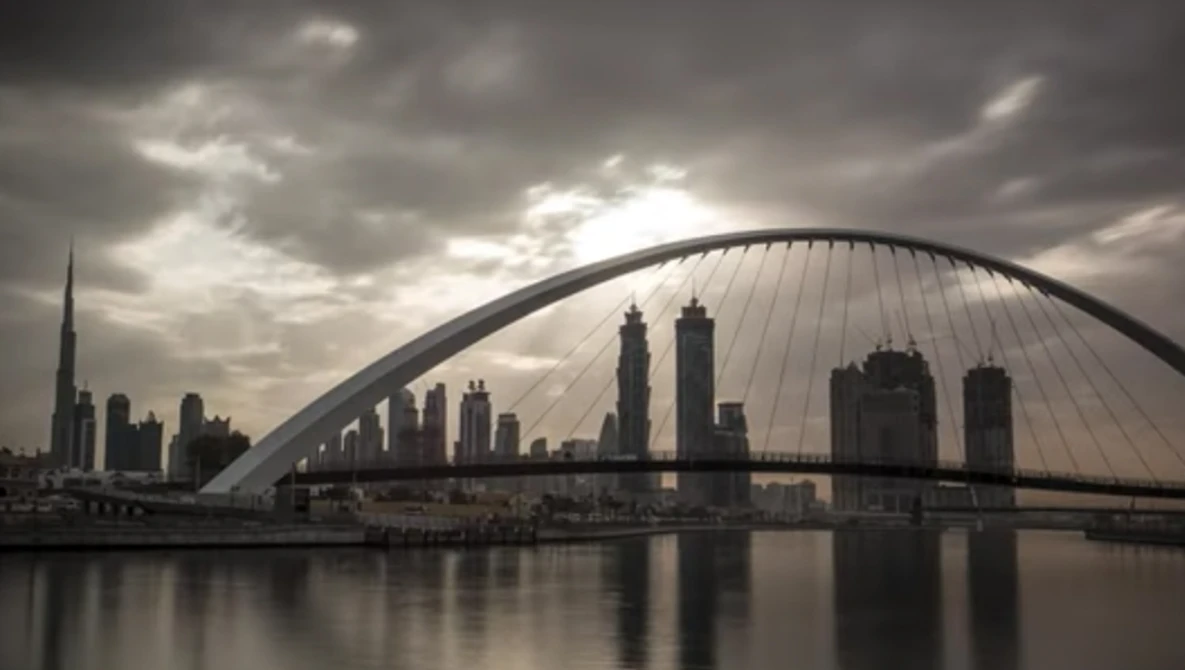 Modern arched bridge spanning a river with city skyline and dramatic cloudy sky reflected in still water.