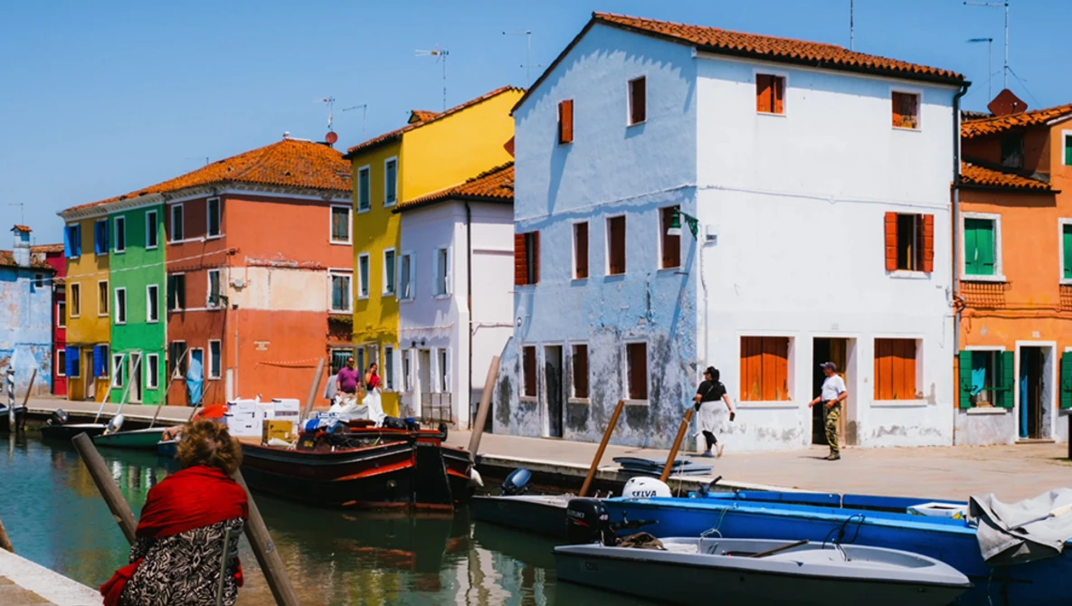 Brightly colored buildings reflected in a canal with moored boats in the foreground.
