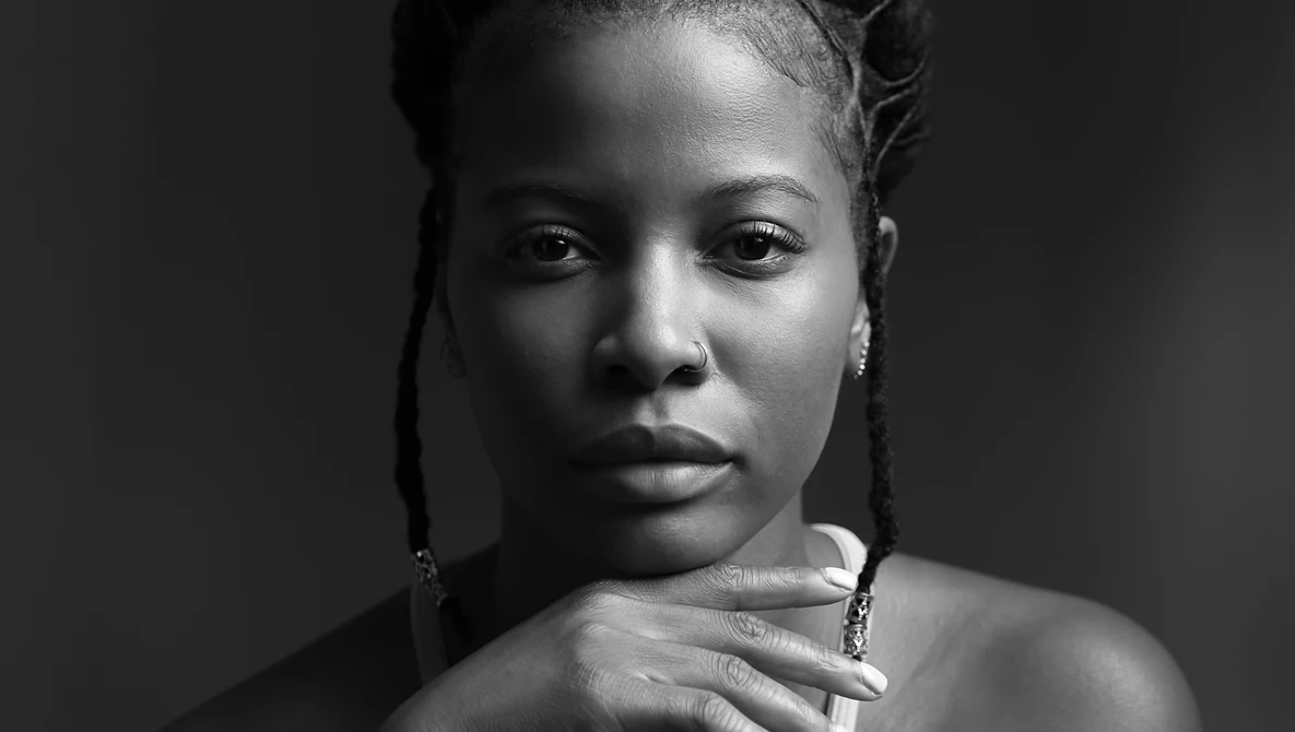 Black and white portrait of a woman with braided hair, looking downward with her hand near her shoulder.