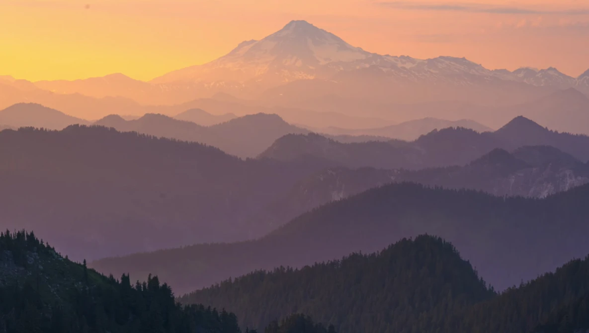 Snow-capped volcanic peak rising above forested mountain ridges at sunrise, with atmospheric haze layering the distant ranges.