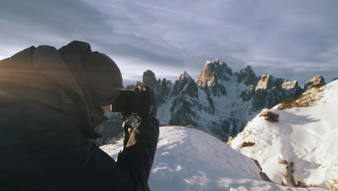 Photographer with camera photographing jagged snow-covered mountain peaks in golden light.