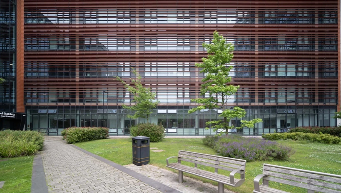 Modern courtyard with wooden benches, manicured lawn, and young trees facing a multi-story building with horizontal wooden slats.