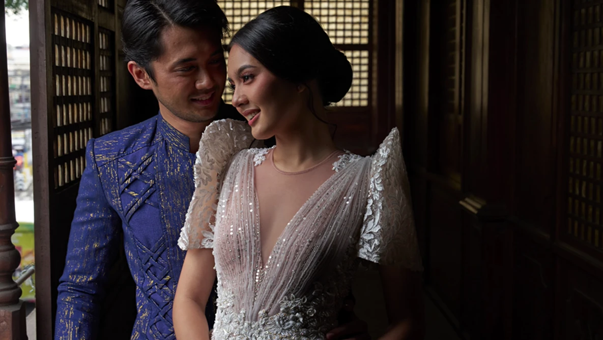 Couple in formal traditional attire standing together indoors with warm architectural lighting.
