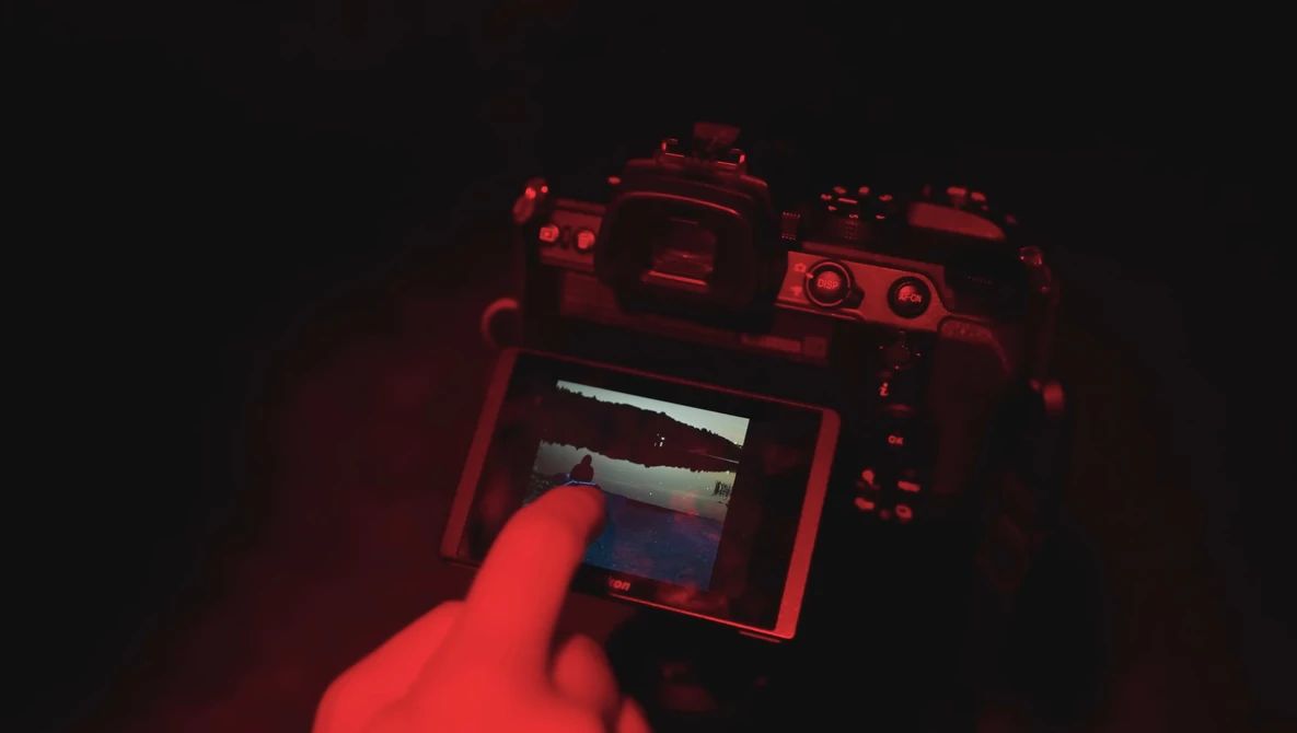 Photographer's hand holding a mirrorless camera with LCD screen visible in red-tinted lighting.