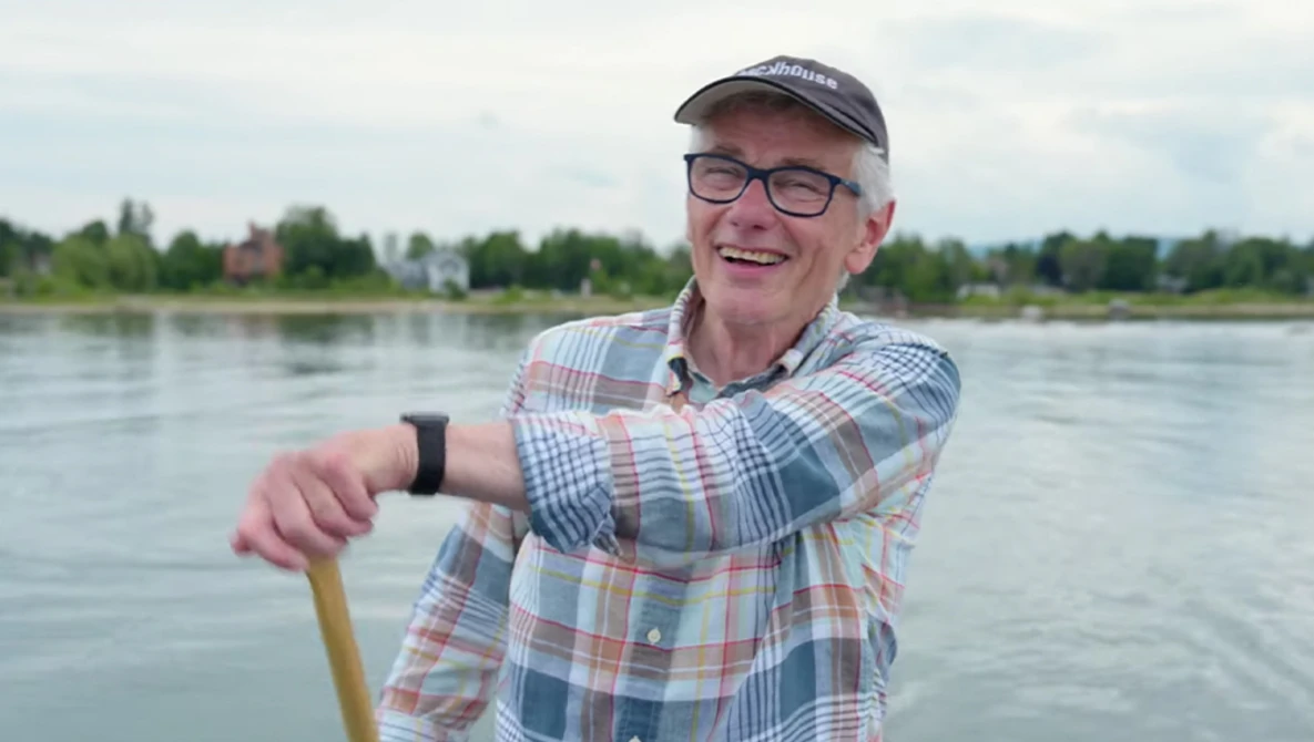 Smiling man in plaid shirt and cap holding paddle by river.