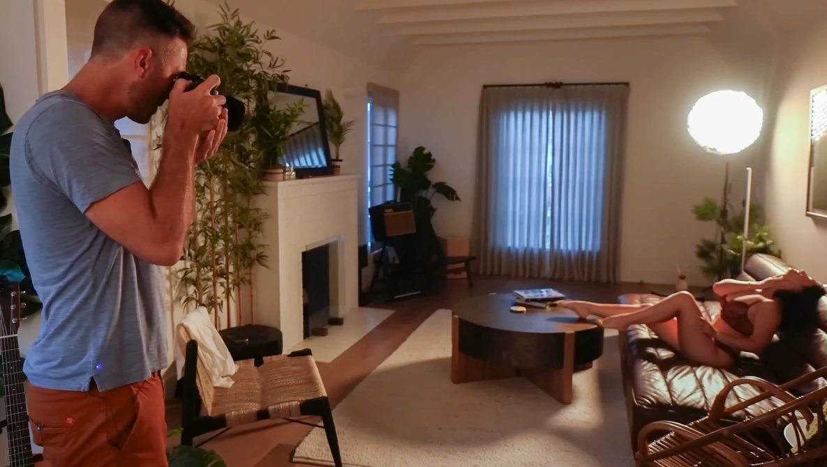 Photographer holding a camera with telephoto lens, composing a shot of a woman reclining on a couch in a modern living room.