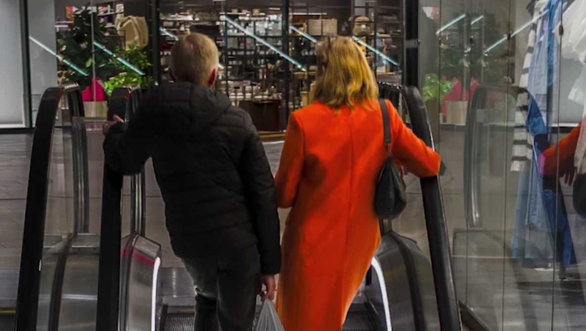Two people viewed from behind riding an escalator toward a storefront with holiday decorations.