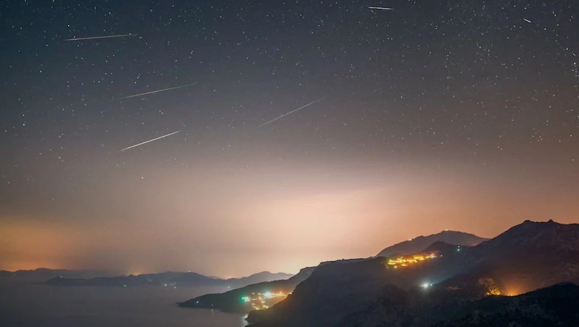 Meteor shower streaking across a twilight sky above a coastal landscape with illuminated hillside settlements.